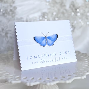 May include: A white card with a scalloped edge, featuring a blue butterfly and the text "SOMETHING BLUE for Beautiful you." A white ribbon is wrapped around the card. The card is displayed on a white, decorative cake stand.