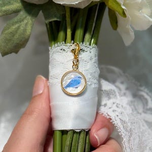 May include: A round gold-toned pendant charm featuring a small blue bird illustration, attached to a white satin ribbon and white lace wrap around a bouquet of white flowers. The charm has a clear circular frame and a gold lobster clasp attachment.