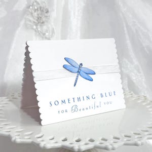 May include: A white card with a scalloped edge, featuring a light blue dragonfly and the text "SOMETHING BLUE FOR Beautiful YOU." A white ribbon accents the card. The card is displayed on a white, decorative plate.
