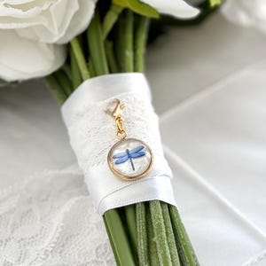 May include: A bridal bouquet featuring white flowers and green stems, wrapped with white lace and ribbon. A gold-colored charm with a blue dragonfly design is attached to the bouquet, adding a delicate touch to the arrangement.