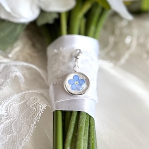 May include: A bouquet of white flowers wrapped with white ribbon and lace. A silver charm with a blue forget-me-not flower is attached to the ribbon. The stems are green.