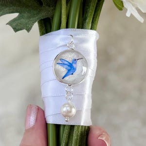 May include: A silver pendant with a blue hummingbird design and a white pearl charm. The pendant is attached to a bouquet of white flowers wrapped with a white ribbon.