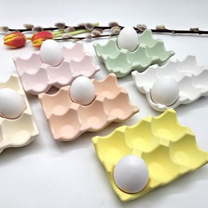 Könnte beinhalten: Eine Sammlung pastellfarbener Eierhalter aus Keramik, die jeweils ein weißes Ei halten. Die Halter sind in den Farbtönen Rosa, Pfirsich, Grün, Weiß und Gelb gehalten. Dekorative Blumen und Zweige im Hintergrund deuten auf ein Frühlingsmotiv hin.