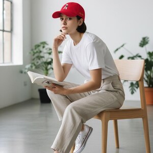Puede incluir: Una gorra de b&eacute;isbol roja con un dise&ntilde;o gr&aacute;fico, una camiseta blanca y pantalones beige. La persona est&aacute; sentada en una silla de madera y leyendo un libro. Zapatillas blancas completan el atuendo.