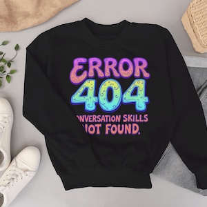 May include: Black sweatshirt with the text "ERROR 404 CONVERSATION SKILLS NOT FOUND" in colourful, bubble-style lettering. The design features a vibrant mix of purple, pink, blue, and yellow. The sweatshirt is paired with jeans and white trainers.
