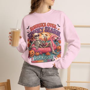 May include: A light pink long-sleeve sweatshirt featuring a graphic print of a skeleton wearing sunglasses, relaxing in a pink flamingo pool float on water. The text reads "SUMMER'S OVER, SPOOKY SEASON LOADING" with pumpkins, bats, and autumn leaves.