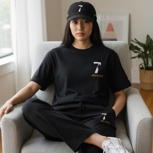 Puede incluir: Conjunto de camiseta, pantal&oacute;n y gorra de b&eacute;isbol negros con un gr&aacute;fico del n&uacute;mero 7 en blanco. La gorra y la camiseta presentan el n&uacute;mero 7 en blanco. Los pantalones tienen el n&uacute;mero 7 en blanco. El modelo lleva zapatillas blancas.