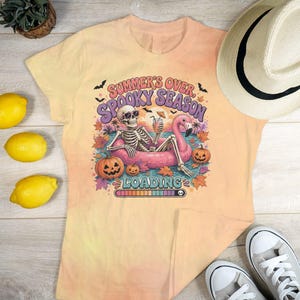 Puede incluir: Una camiseta color melocotón con un gráfico de un esqueleto con gafas de sol descansando sobre un flotador de flamenco rosa. El texto dice "SUMMER'S OVER, SPOOKY SEASON LOADING" con una barra de carga, calabazas, murciélagos y hojas de otoño.