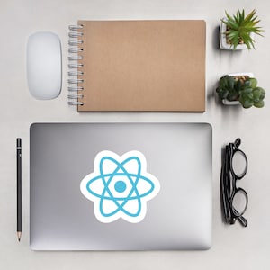 May include: A silver laptop computer with a blue and white React logo sticker on the lid. The laptop is on a white surface with a notebook, a mouse, a pencil, glasses, and two small potted plants.