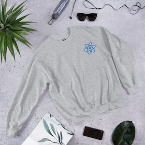 Peut inclure: Un sweat-shirt gris avec un symbole d'atome bleu sur la poitrine gauche. Le sweat-shirt est posé à plat sur une surface grise.