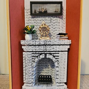 Può includere: Un'opera d'arte di libro fatta a mano con una scena di camino. Il camino è realizzato con pagine di libro piegate, con un dipinto in miniatura, un orologio e una pianta. Le pagine del libro sono visibili, creando un effetto strutturato, con accenti rossi e gialli.