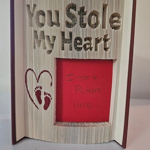 Op de afbeelding: Een boek met de woorden "You Stole My Heart" uitgeknipt uit de pagina's. Het boek is open om een rode kaart te laten zien met de woorden "Insert Picture Here" erop geschreven. Een hart met voetafdrukken is uitgeknipt uit de pagina links van de kaart.