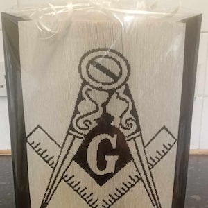 Könnte beinhalten: Eine schwarz-weiße Papierkunst-Skulptur eines Freimaurer-Symbols, mit einem Zirkel und einem Winkelmaß mit einem 'G' in der Mitte. Die Skulptur ist in durchsichtige Plastikfolie verpackt und hat einen schwarzen Rahmen.