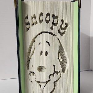 May include: A book with the pages folded to create a 3D sculpture of Snoopy, the Peanuts character. The sculpture is painted in black and white and the book is bound in blue with green endpapers.