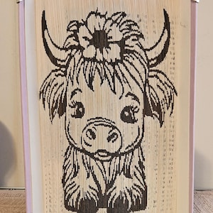 Puede incluir: Una pieza de arte de libro única con un diseño de vaca de las Tierras Altas. La ilustración en blanco y negro de la vaca se crea doblando las páginas de un libro. Los bordes están protegidos con protectores de esquina dorados y el lomo es lavanda.
