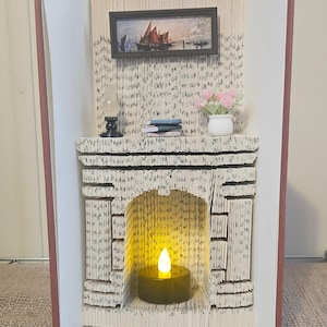 Op de afbeelding: Een miniatuur haardsculptuur gemaakt van gevouwen pagina's uit een boek. De haard heeft een ingelijst schilderij, een kleine lamp, boeken en een vaas met roze bloemen. Een batterij-aangedreven kaars gloeit in de haard en creëert een warme sfeer.
