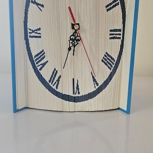 Op de afbeelding: Een boek gevouwen in de vorm van een klok met Romeinse cijfers. De wijzerplaat is zwart met witte Romeinse cijfers en de wijzers zijn zwart en rood.