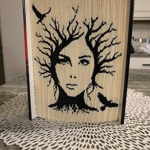 May include: Folded book art depicting a woman's face with tree-like hair and two black birds. The image is created by folding the pages of a book, forming a unique artistic design. The book is standing upright.