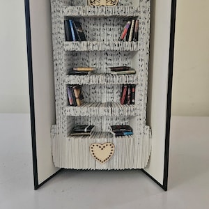 Könnte beinhalten: Ein Bücherregal in Buchform mit fünf Regalbrettern, die jeweils mit kleinen Büchern gefüllt sind. Das Bücherregal besteht aus gefalteten Seiten eines Buches und hat unten ein herzförmiges Design. Das Bücherregal ist offen und steht auf einer weißen Oberfläche.