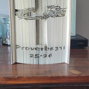 Könnte beinhalten: Ein Buch mit Seiten, die zu einem 3D-Kreuz gefaltet sind. Das Kreuz ist mit einer Dornenkrone verziert. Das Buch trägt den Titel "Sprüche 3:13" und "25:26".