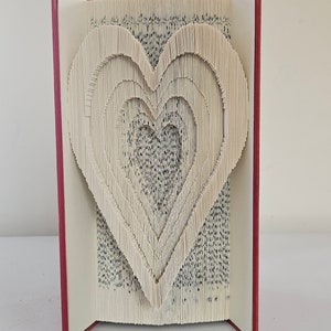 May include: A book with pages folded into a three-dimensional heart shape. The book is open and the heart is visible. The book has a red cover.