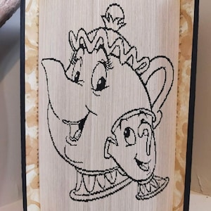 May include: A black and white book folding art piece depicting Mrs. Potts and Chip from Disney's Beauty and the Beast.