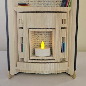 Peut inclure: Un livre plié en forme de cheminée avec une petite bougie à l'intérieur. La cheminée a une étagère sur le dessus et deux étagères sur les côtés. Le livre est décoré d'accents dorés.