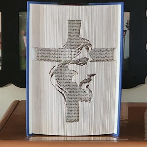 May include: A book with a cross design carved into the pages. The pages are folded to create the image of a cross with a bird. The book has blue covers and is displayed on a wooden surface.