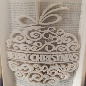 May include: A white book with the pages folded into a three-dimensional design of a Christmas ornament. The ornament has a swirl pattern and the words "Merry Christmas" are written on it.