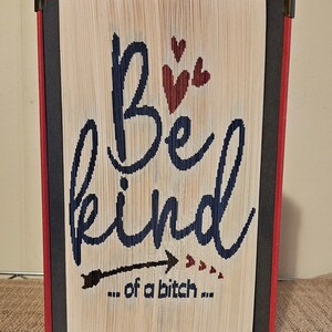 Op de afbeelding: Gevouwen boekkunst met de tekst "Be kind... of a bitch..." in marineblauw. Rode harten en pijlaccenten zijn inbegrepen. Het boek heeft rode en zwarte randen met metalen hoeken.