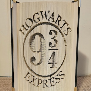 May include: A book sculpture with the words "HOGWARTS 9 3/4 EXPRESS" carved into the pages. The book is open, with the carved design visible. The edges of the book are black, with metal corner protectors.
