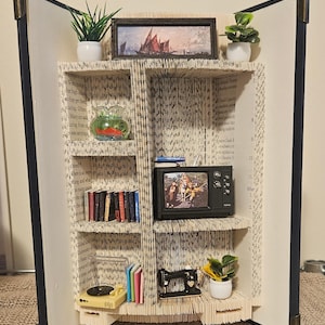 Può includere: Una libreria in miniatura realizzata con le pagine di un libro. La libreria ha quattro ripiani ed è decorata con un piccolo televisore, un giradischi, una macchina da cucire, una boccia di pesce e due piante in vaso. La libreria è aperta e le pagine del libro sono visibili.