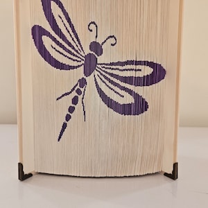 May include: A book art sculpture of a purple dragonfly, folded from the pages of a book and mounted on a wooden base with metal corner accents.