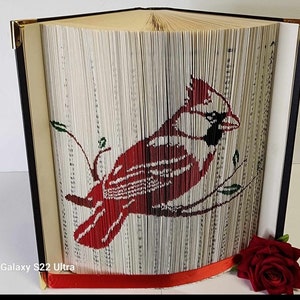 Op de afbeelding: Een boekkunst sculptuur van een rode kardinaal die op een tak zit. Het boek is gevouwen en gevormd om de afbeelding van de vogel te creëren. Het beeld wordt op een tafel met een rode roos tentoongesteld.