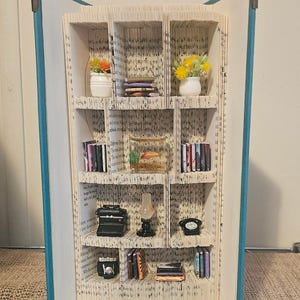 Könnte beinhalten: Eine Buchnischen-Skulptur aus einem gefalteten Buch mit blauem Einband. Das Innere zeigt Miniaturregale mit winzigen Büchern, einem Goldfischglas, einer schwarzen Schreibmaschine, einer kleinen Lampe, einem Telefon, einer Kamera und zwei Blumentöpfen.