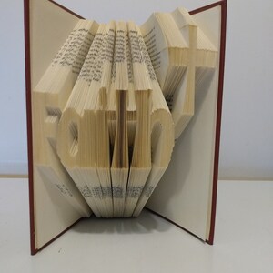 Puede incluir: Un libro con la palabra "Fe" doblada en las páginas. El libro está abierto y las páginas están dobladas para crear un efecto 3D. El libro tiene una cubierta roja.