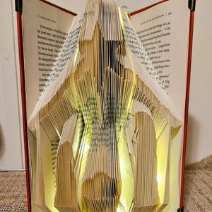 May include: A folded book sculpture, illuminated by warm LED lights, forming a star shape. The pages are intricately folded to create an architectural design. The book has a red cover with black corner protectors. A unique decorative item.