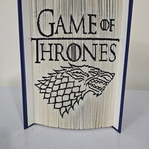 Peut inclure: Un livre avec les mots "Game of Thrones" sur la couverture, avec une illustration en noir et blanc d'un loup-garou sur la face avant.