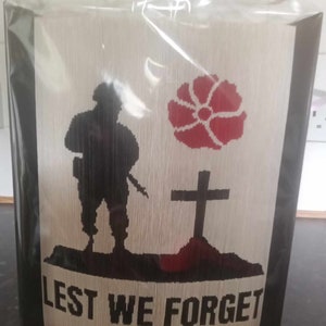 Op de afbeelding: Een gevouwen boekkunstwerk met een zwart silhouet van een soldaat die op een heuvel staat met een rode klaproos en een kruis. De tekst "LEST WE FORGET" is onder de afbeelding gedrukt.