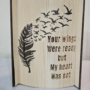 Puede incluir: Una escultura de arte de libro con una pluma negra y una bandada de pájaros volando. El texto dice "Tus alas estaban listas pero mi corazón no lo estaba."