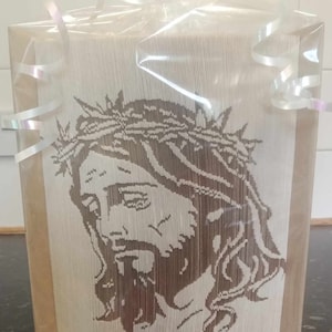 Peut inclure: Une boîte cadeau en papier brun avec un ruban blanc et un couvercle en plastique transparent. La boîte présente une image en papier de Jésus avec une couronne d'épines.