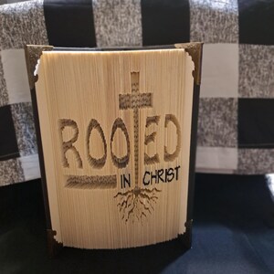 May include: A handcrafted book art piece featuring the inscription "ROOTED IN CHRIST" carved into the pages. The design includes a cross and roots, with brass corner protectors. The book is set against a black and white checked backdrop.