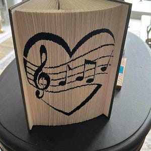 May include: An open book with pages folded to create a black heart design featuring musical notes and a treble clef. The book is displayed on a dark table. The design is visible on the pages.