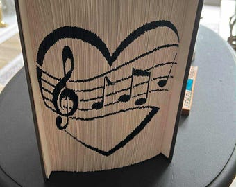 May include: An open book with pages folded to create a black heart design featuring musical notes and a treble clef. The book is displayed on a dark table. The design is visible on the pages.