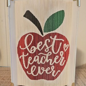 May include: A handmade book art piece featuring a red apple design with a green leaf and the words "Best Teacher Ever" in white script. The artwork is made from folded book pages and has gold corner protectors.