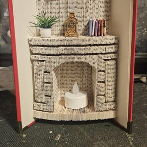Peut inclure: Une sculpture de cheminée unique et artisanale, réalisée à partir d'un livre recyclé. Comprend une peinture miniature, une horloge, une plante et une bougie. La cheminée est faite de pages pliées, créant un effet texturé.