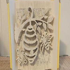 May include: A folded book art sculpture featuring a central beehive hanging from a branch with two bees on either side. The book has light yellow covers with metallic corner protectors, and the pages are intricately folded to create the design.