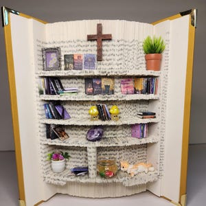 May include: A book-shaped bookshelf with a cross on top, filled with miniature books, plants, and figurines. The bookshelf is made of folded pages from a book.