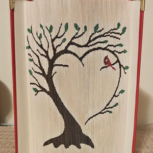 May include: A book art sculpture of a tree with green leaves forming a heart shape with a red cardinal perched on a branch. The book is bound in red with gold corner accents.