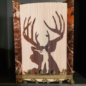 Peut inclure: Une sculpture d'art de livre représentant un cerf avec de grands bois, réalisée à partir de pages d'un livre. La sculpture est décorée d'un tissu camouflage et d'un ruban vert avec des accents de feuilles.